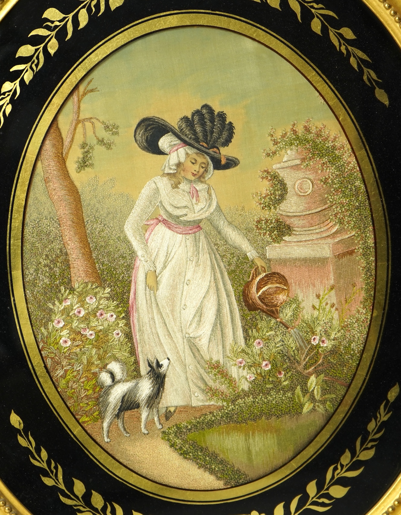A pair of early 20th century oval silk embroideries, one of a mother and child dancing in a garden, the other of a lady in a large brimmed hat, watering flowers in by a classical stone urn, watched by her dog. Framed in